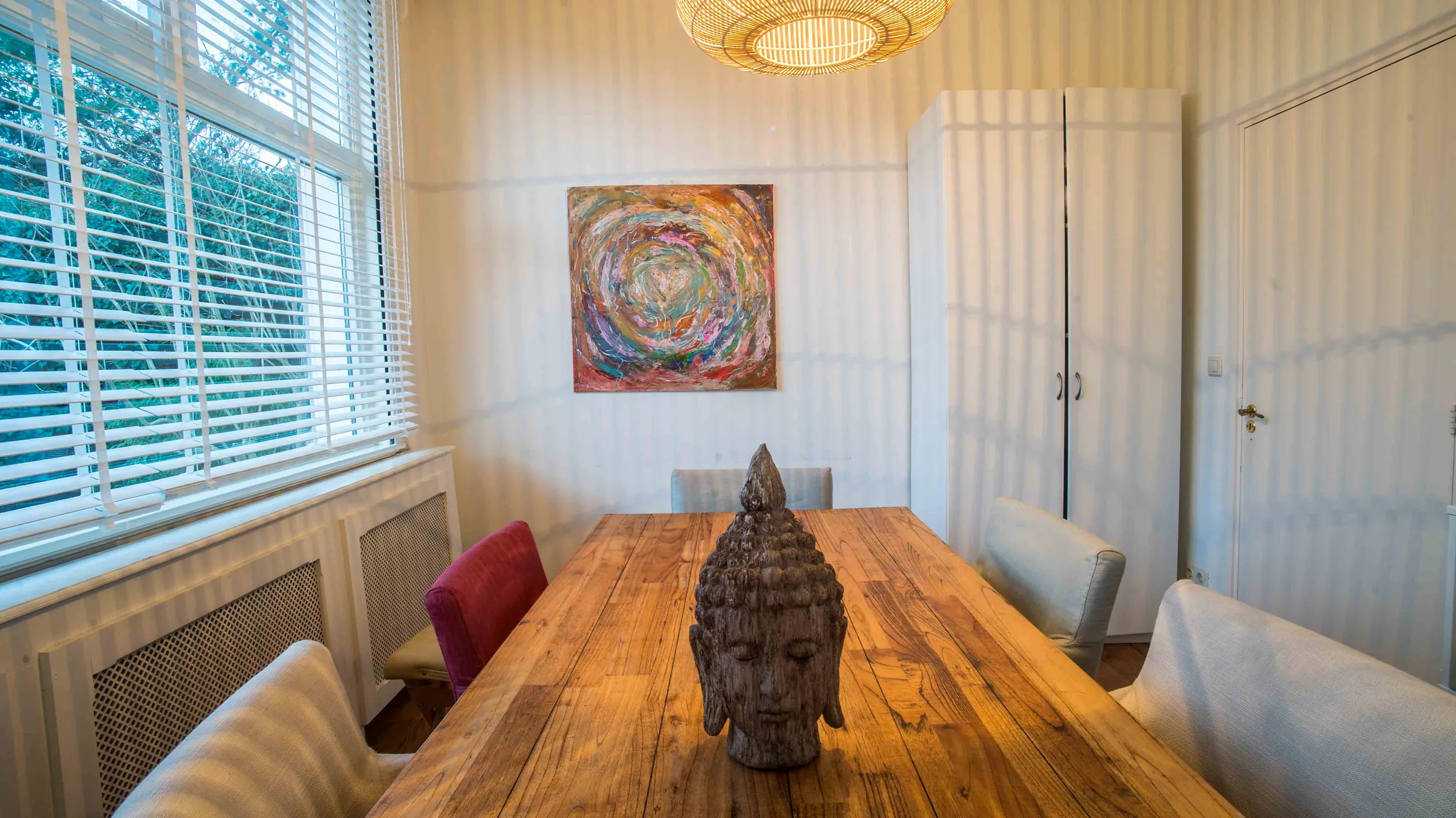 De coachings- en vergaderruimte: een massieve houten tafel met stoelen eromheen, een houten Boeddhahoofd op tafel en daglicht door de jaloezieën.
