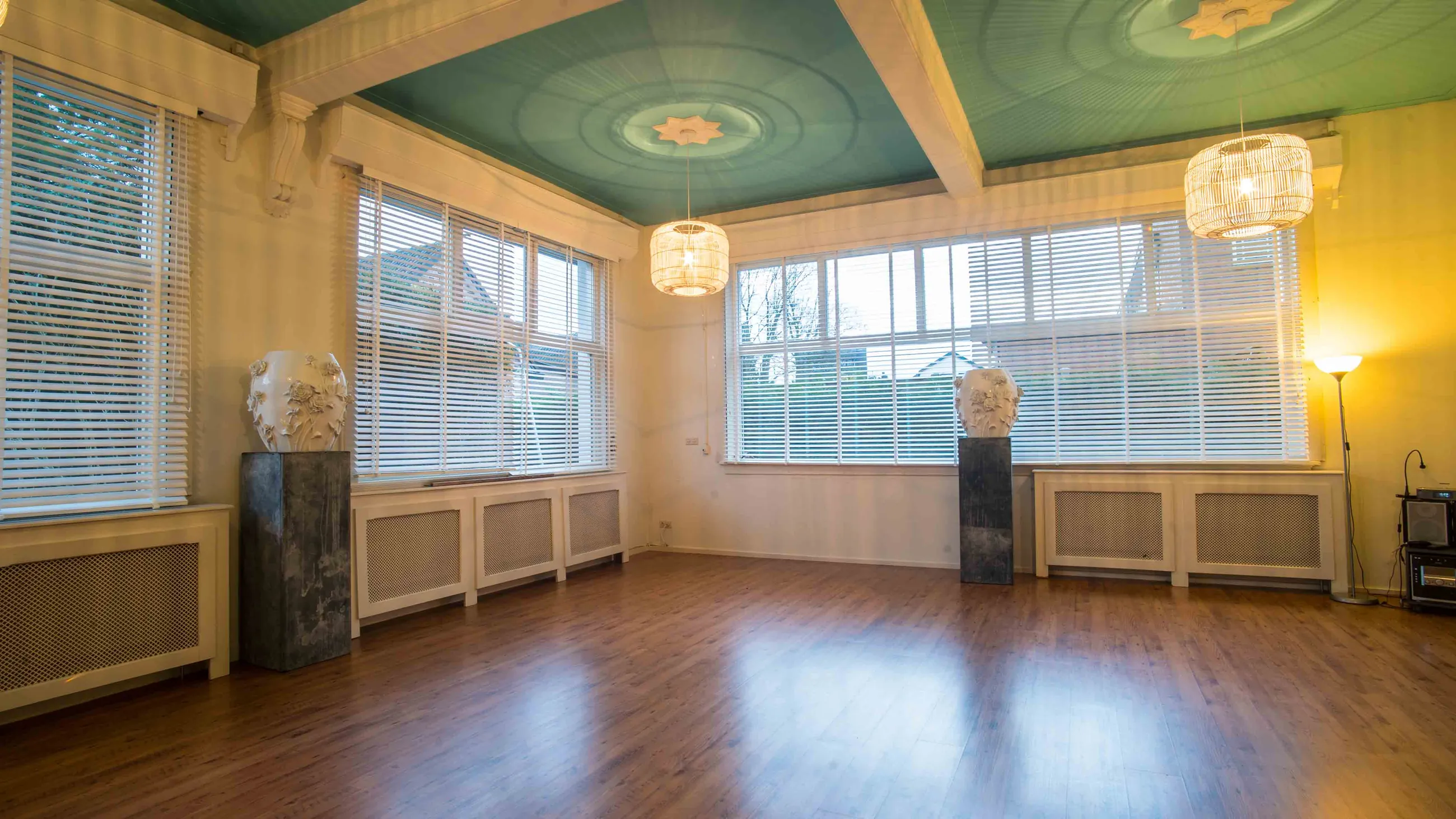 De grote presentatiezaal: een lege houten vloer, witte wanden en het turkooise plafond met geschilderde rozetten.